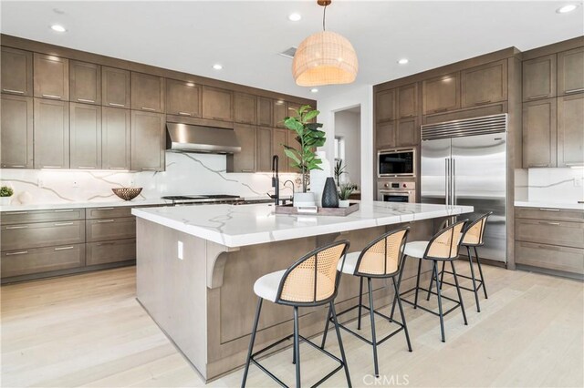 kitchen featuring extractor fan, hanging light fixtures, built in appliances, a center island with sink, and light hardwood / wood-style flooring