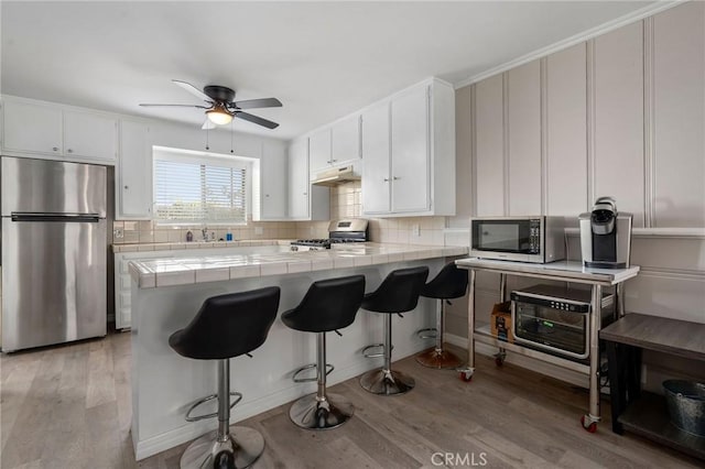 kitchen featuring backsplash, tile counters, under cabinet range hood, appliances with stainless steel finishes, and light wood-style floors