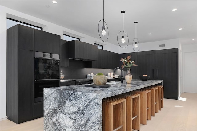 kitchen featuring pendant lighting, double oven, dark stone counters, a center island with sink, and wall chimney range hood