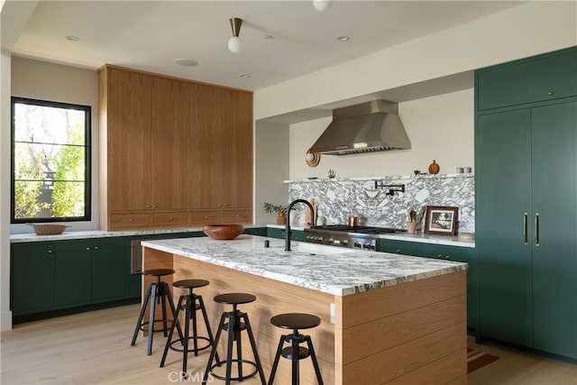 kitchen featuring light hardwood / wood-style floors, a kitchen breakfast bar, an island with sink, wall chimney exhaust hood, and light stone counters