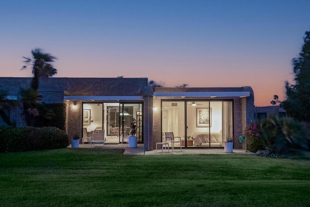 back house at dusk featuring a lawn and a patio