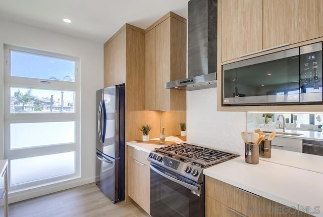 kitchen featuring appliances with stainless steel finishes, wall chimney range hood, light brown cabinetry, sink, and light wood-type flooring