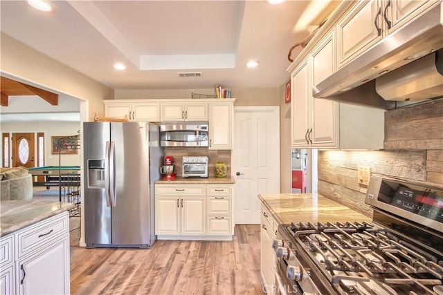 kitchen featuring appliances with stainless steel finishes, backsplash, a raised ceiling, light wood-type flooring, and light stone countertops