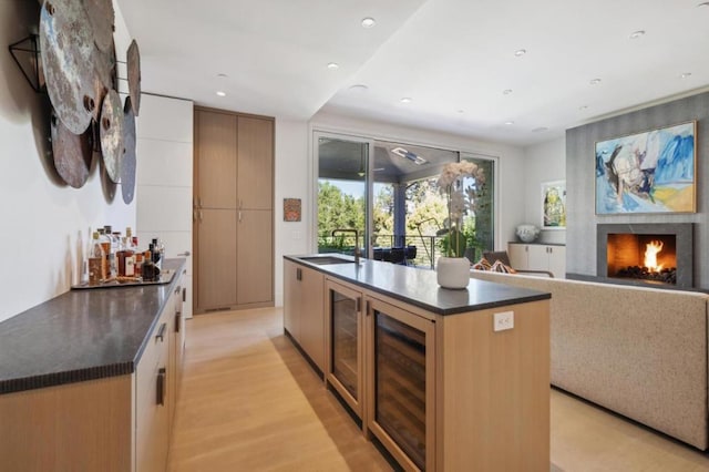 kitchen with a large fireplace, sink, light wood-type flooring, a kitchen island with sink, and beverage cooler