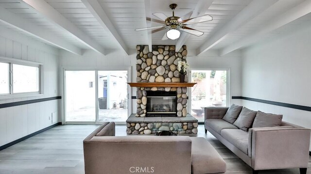 living area featuring a stone fireplace, beamed ceiling, a ceiling fan, and wood finished floors