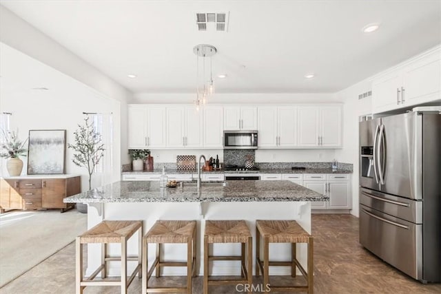 kitchen with dark stone countertops, white cabinetry, hanging light fixtures, an island with sink, and stainless steel appliances