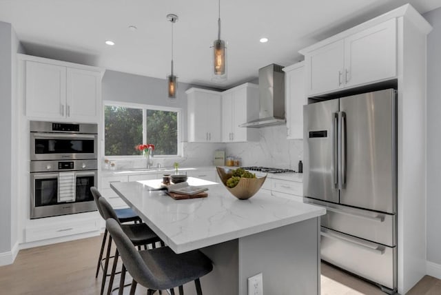 kitchen featuring wall chimney exhaust hood, white cabinetry, hanging light fixtures, appliances with stainless steel finishes, and a kitchen island