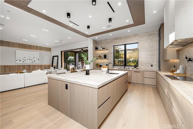 kitchen featuring a large island with sink, wall chimney range hood, sink, light brown cabinets, and black electric cooktop