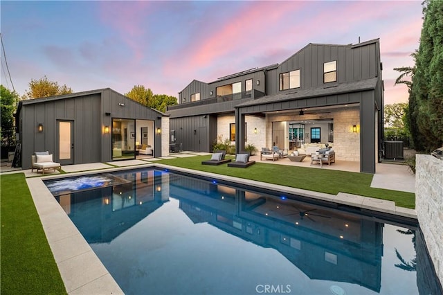 back house at dusk featuring central AC unit, an outbuilding, outdoor lounge area, ceiling fan, and a pool with hot tub