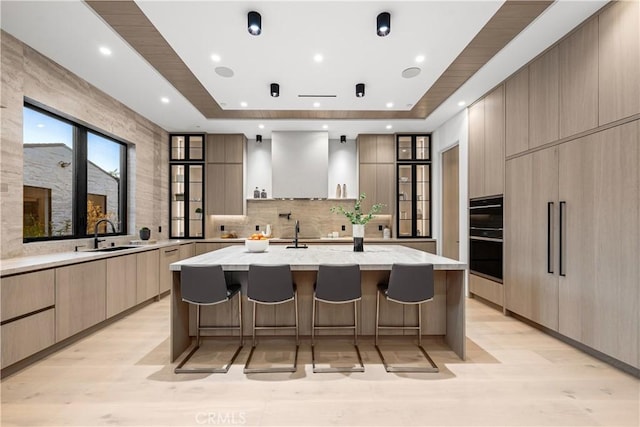 kitchen with a spacious island, light stone countertops, a kitchen bar, wall chimney range hood, and sink