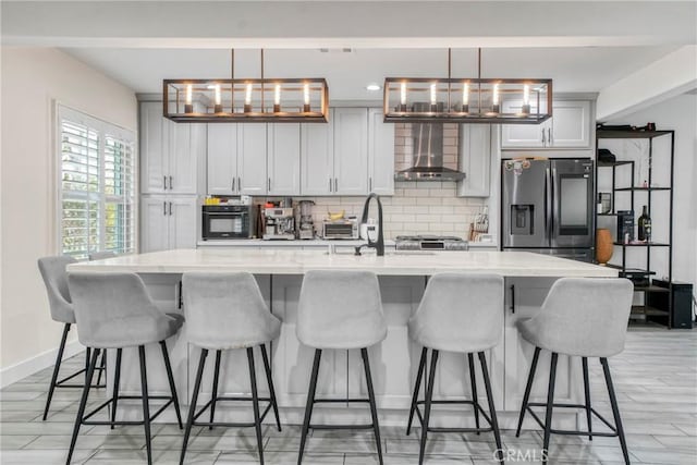 kitchen with baseboards, a kitchen island with sink, stainless steel fridge, wall chimney range hood, and tasteful backsplash