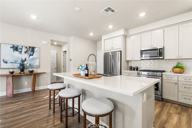 kitchen featuring appliances with stainless steel finishes, dark hardwood / wood-style floors, an island with sink, a kitchen bar, and decorative backsplash