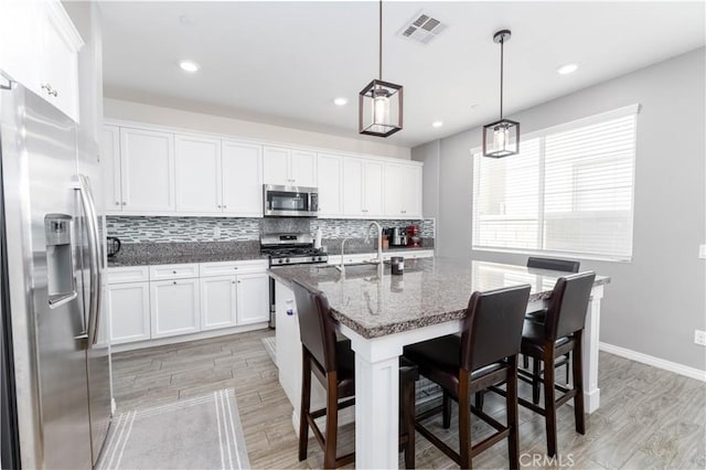 kitchen with dark stone countertops, a center island with sink, hanging light fixtures, appliances with stainless steel finishes, and white cabinets