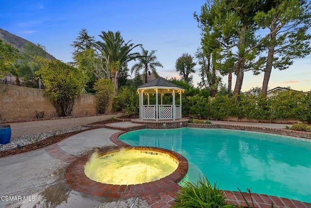 pool at dusk with a gazebo and an in ground hot tub