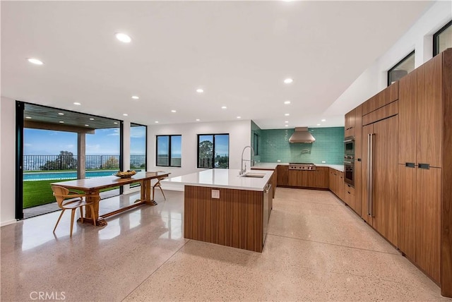 kitchen with wall oven, a kitchen island with sink, gas stovetop, wall chimney exhaust hood, and sink