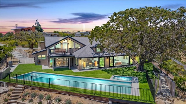 back house at dusk with a swimming pool with hot tub, a balcony, and a yard