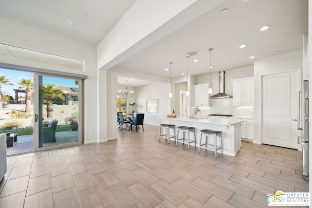 kitchen featuring pendant lighting, white cabinets, wall chimney range hood, an island with sink, and a breakfast bar