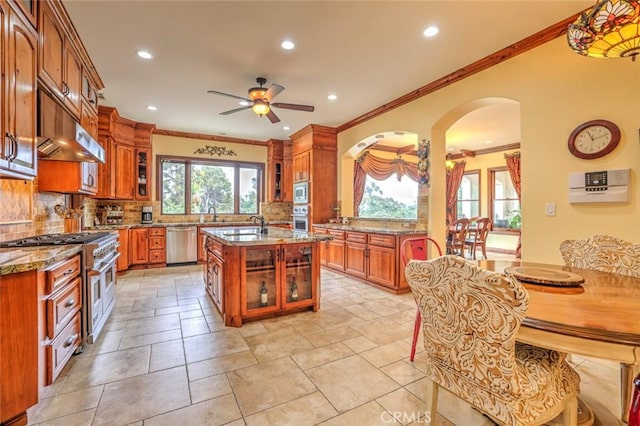 kitchen featuring appliances with stainless steel finishes, a kitchen island with sink, ceiling fan, light stone counters, and crown molding