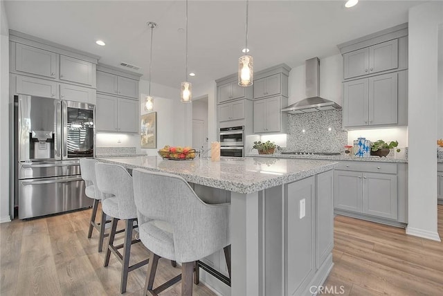 kitchen featuring hanging light fixtures, wall chimney exhaust hood, an island with sink, light hardwood / wood-style floors, and stainless steel appliances