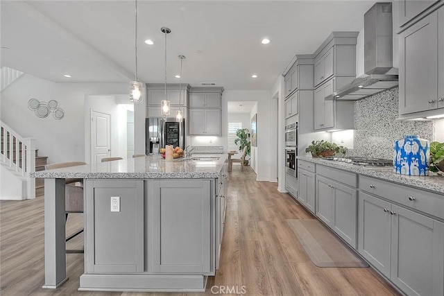 kitchen featuring wall chimney range hood, a kitchen island with sink, gray cabinetry, and a kitchen breakfast bar