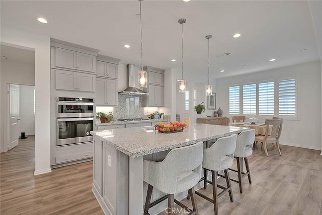 kitchen featuring decorative light fixtures, wall chimney range hood, stainless steel appliances, a breakfast bar, and a center island with sink