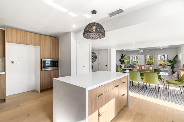 kitchen with decorative light fixtures, ceiling fan, stainless steel microwave, a kitchen island, and light hardwood / wood-style flooring