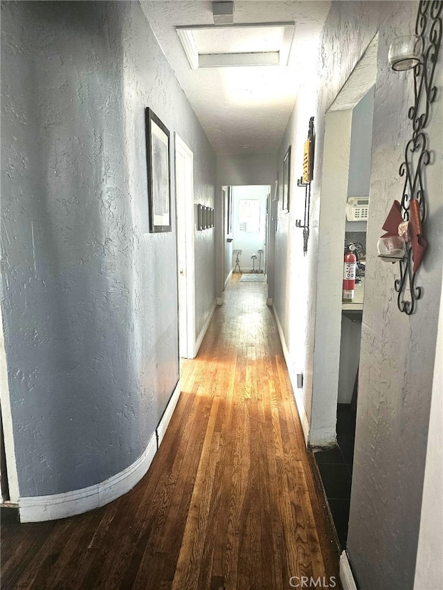 corridor with a textured wall, wood finished floors, attic access, and baseboards