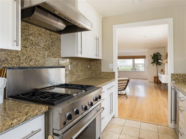 kitchen featuring stone counters, stainless steel gas stove, white cabinets, decorative backsplash, and wall chimney range hood