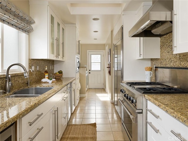 kitchen with sink, white cabinetry, built in appliances, light tile patterned floors, and wall chimney range hood