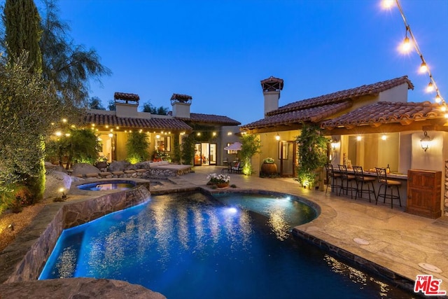 pool at dusk with a patio area, an in ground hot tub, and exterior bar