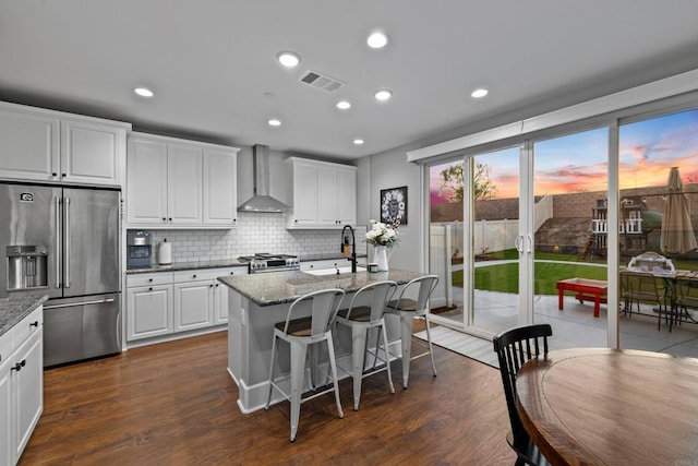 kitchen with a center island with sink, wall chimney range hood, white cabinets, stainless steel appliances, and backsplash