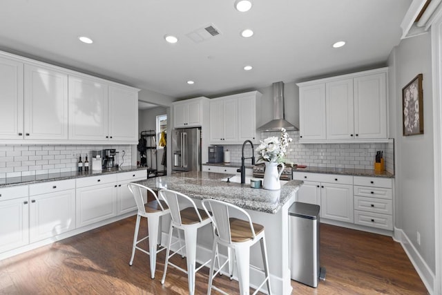 kitchen featuring an island with sink, high end fridge, wall chimney range hood, and white cabinets