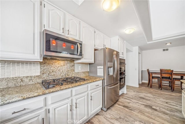 kitchen with light stone countertops, appliances with stainless steel finishes, white cabinetry, light wood-type flooring, and backsplash