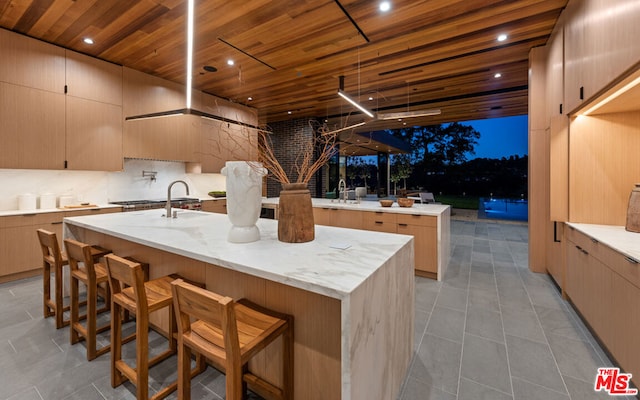 kitchen featuring a kitchen island with sink, wood ceiling, a kitchen breakfast bar, and light brown cabinetry