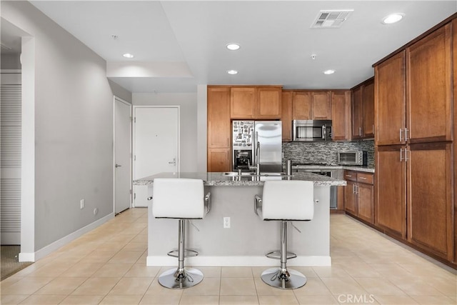 kitchen with appliances with stainless steel finishes, tasteful backsplash, a kitchen island with sink, a kitchen breakfast bar, and light stone countertops