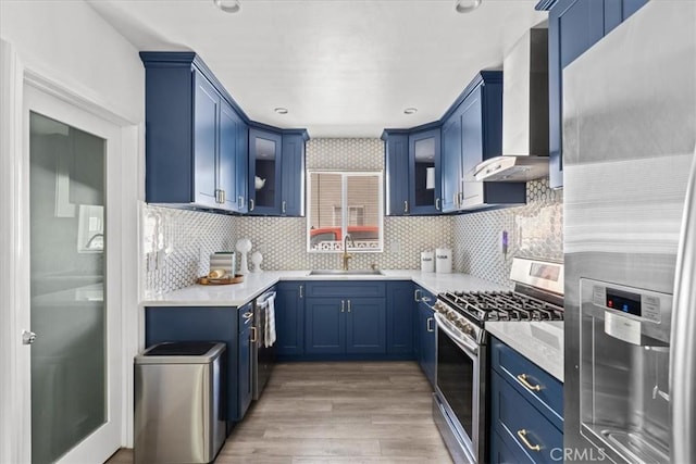 kitchen with wall chimney range hood, stainless steel appliances, sink, backsplash, and blue cabinetry