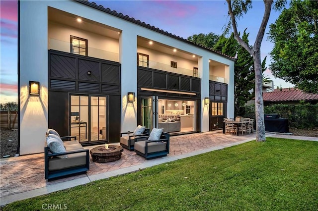 back house at dusk featuring a yard, a patio area, a balcony, and an outdoor fire pit