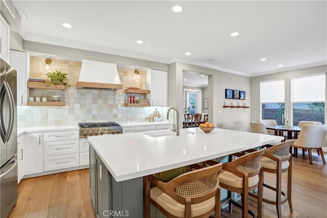 kitchen featuring sink, premium range hood, tasteful backsplash, white cabinets, and a center island with sink