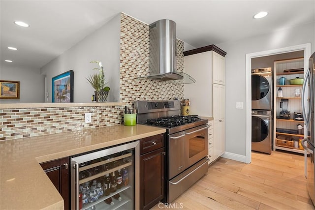 kitchen featuring wall chimney range hood, stacked washer and dryer, backsplash, stainless steel appliances, and wine cooler