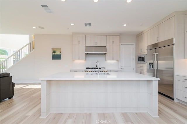 kitchen with a kitchen island with sink, built in appliances, light hardwood / wood-style floors, and range hood