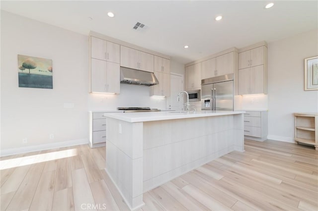 kitchen with sink, range hood, built in appliances, a center island with sink, and light wood-type flooring