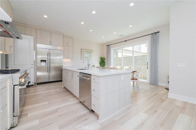 kitchen with sink, high quality appliances, light hardwood / wood-style floors, a center island with sink, and exhaust hood