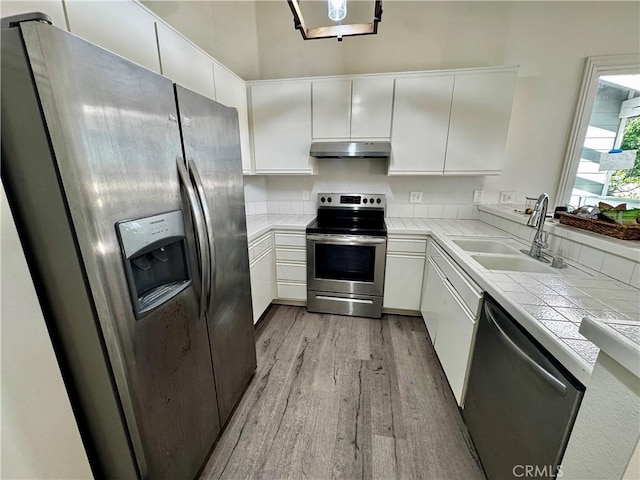 kitchen with under cabinet range hood, a sink, white cabinets, appliances with stainless steel finishes, and tile counters