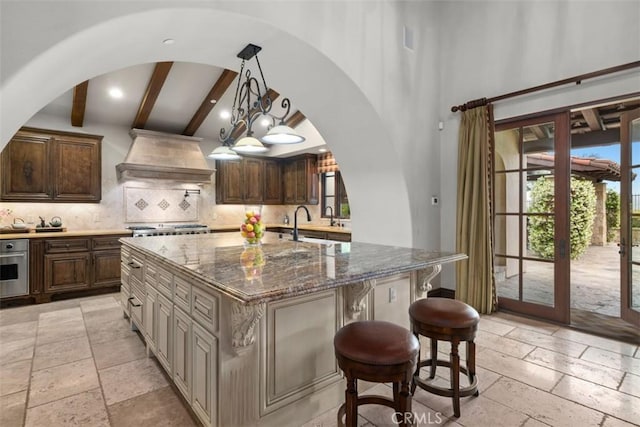 kitchen with backsplash, a large island with sink, dark stone countertops, beam ceiling, and a kitchen bar