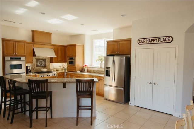 kitchen featuring a breakfast bar area, dark stone counters, a kitchen island with sink, exhaust hood, and stainless steel refrigerator with ice dispenser