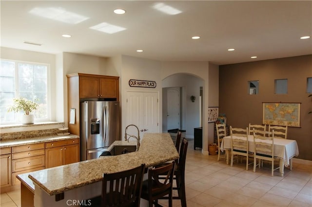 kitchen featuring stainless steel refrigerator with ice dispenser, light tile patterned flooring, light stone countertops, and a kitchen island with sink