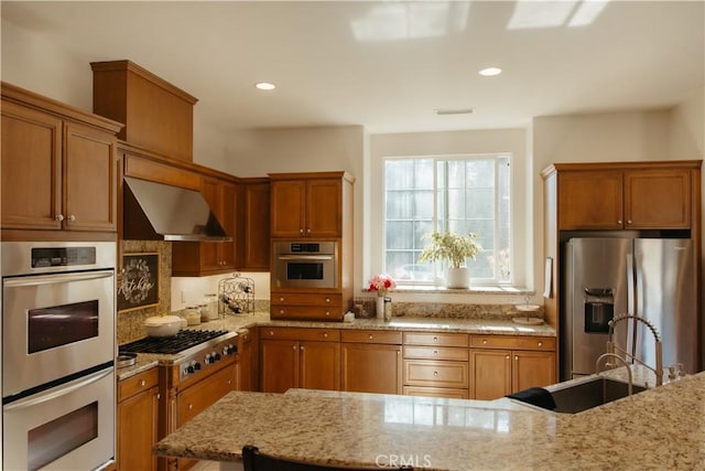 kitchen with stainless steel appliances, light stone countertops, sink, and wall chimney range hood