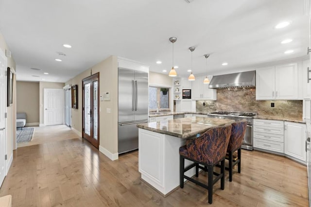 kitchen with wall chimney range hood, white cabinetry, a center island, high end appliances, and light stone counters