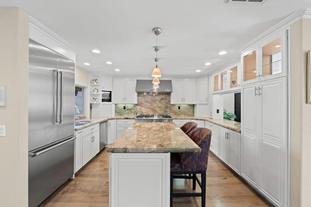 kitchen featuring wall chimney exhaust hood, white cabinetry, decorative light fixtures, appliances with stainless steel finishes, and a kitchen island