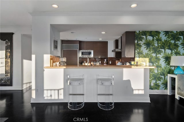 kitchen featuring wall chimney range hood, backsplash, built in appliances, dark brown cabinetry, and ornamental molding